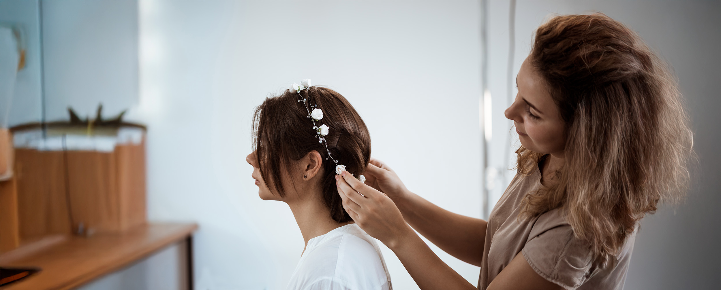 Hairstylist placing a white floral and crystal hair vine into a bride's brown hair.