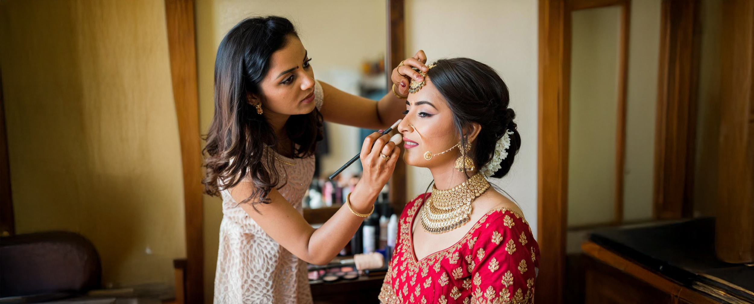 Makeup artist applying blush to an Indian bride in a red lehenga with heavy gold bridal jewelry.
