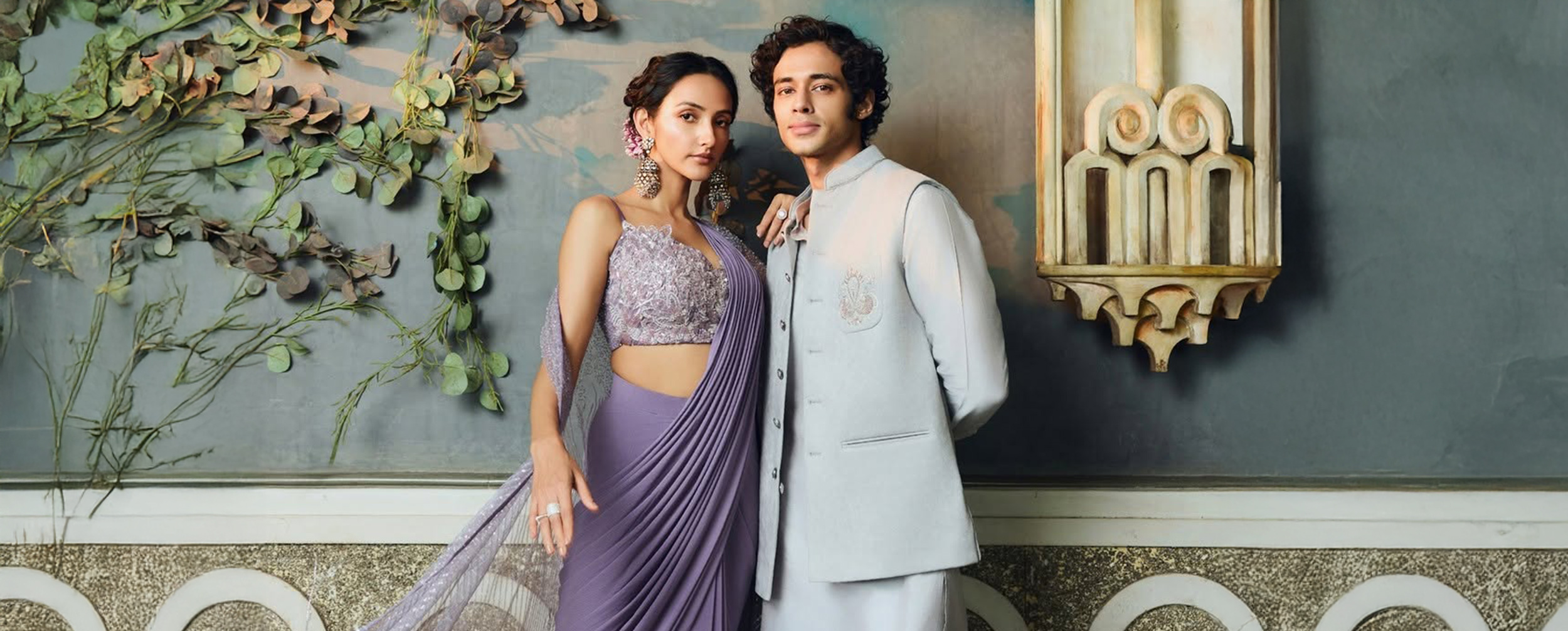 Couple modeling a lavender pre-draped saree and a light grey kurta with Nehru jacket, showcasing fabric and style for Indian festive wear.