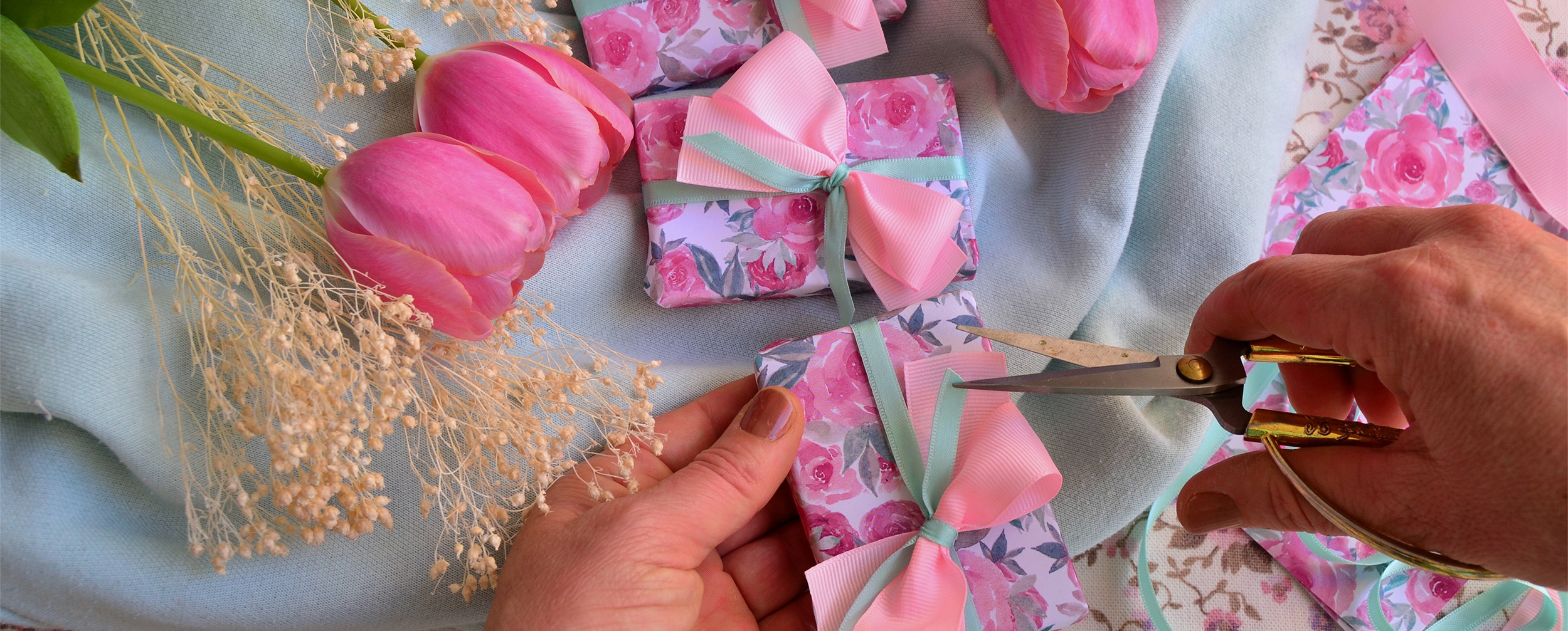 Hands wrapping small wedding gifts in pink floral paper with mint green and pink silk bows, surrounded by pink tulips.