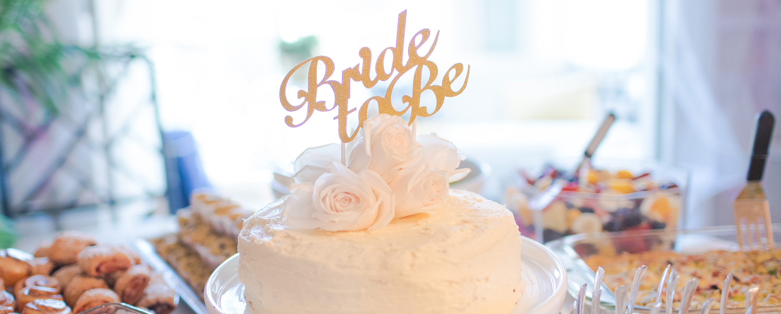 Small white celebration cake topped with blush roses and a gold 'Bride to Be' script topper, surrounded by snacks.
