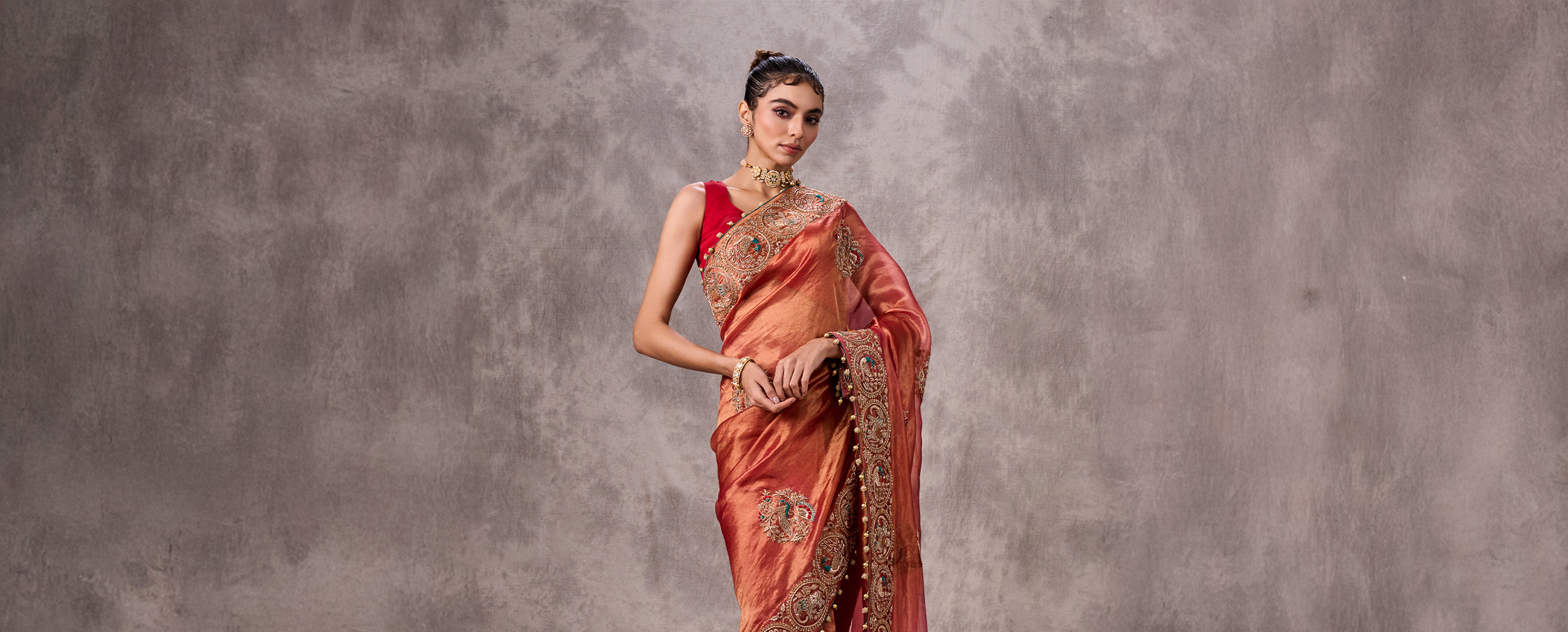 Woman in a rust-orange sheer saree with elaborate embroidery and a contrasting red sleeveless blouse.