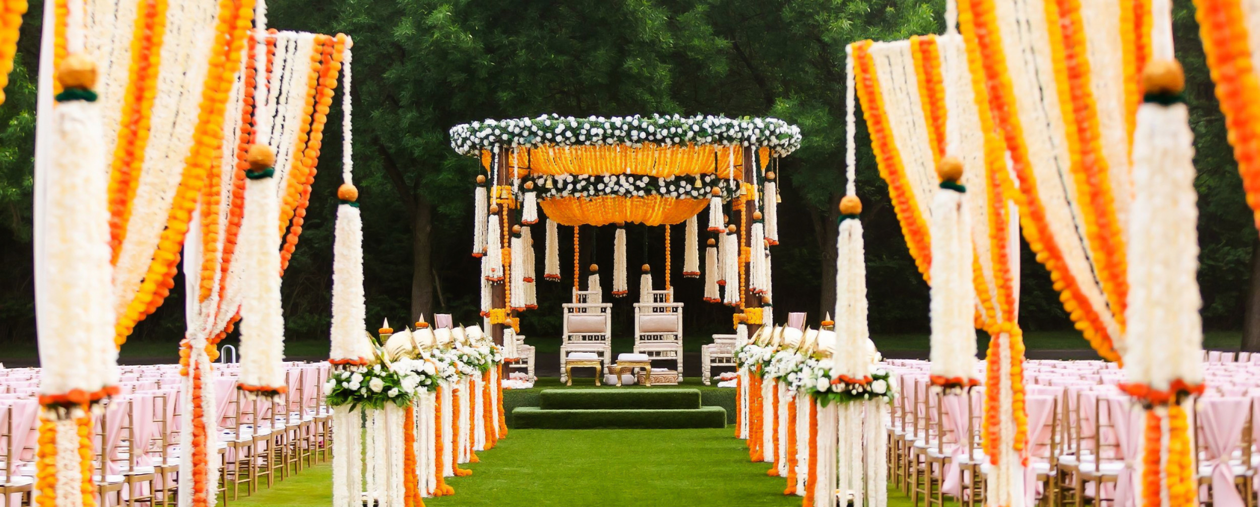 Regal outdoor wedding mandap decorated with dense marigold and jasmine garlands on a green lawn with a flower-lined aisle.