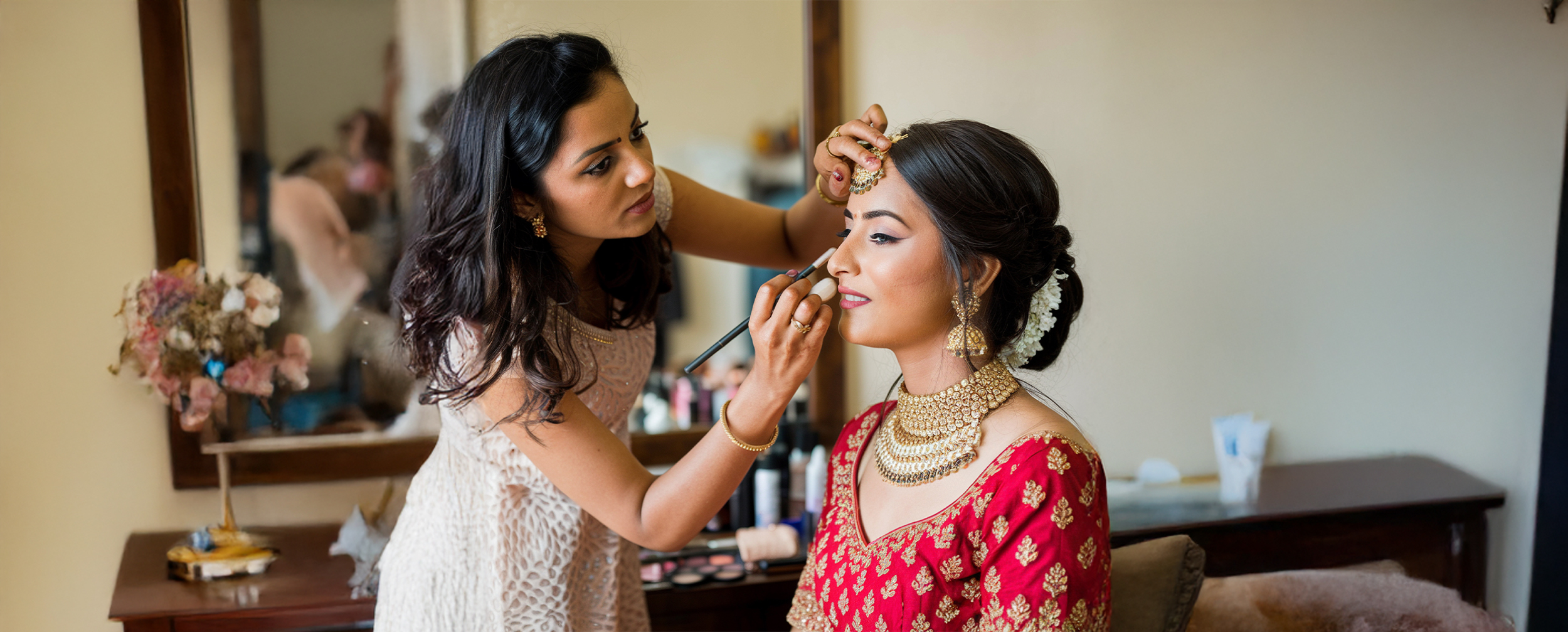 Makeup artist applying concealer to an Indian bride in a red lehenga with traditional gold jewelry.