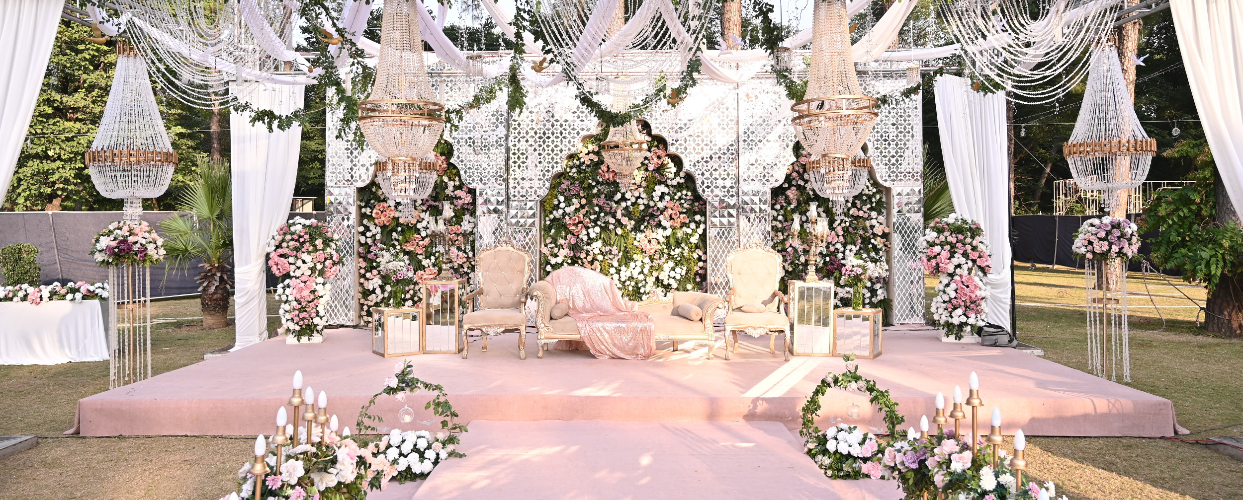 Opulent pink and white floral wedding stage with elaborate lattice backdrop, crystal chandeliers, and white tufted furniture.
