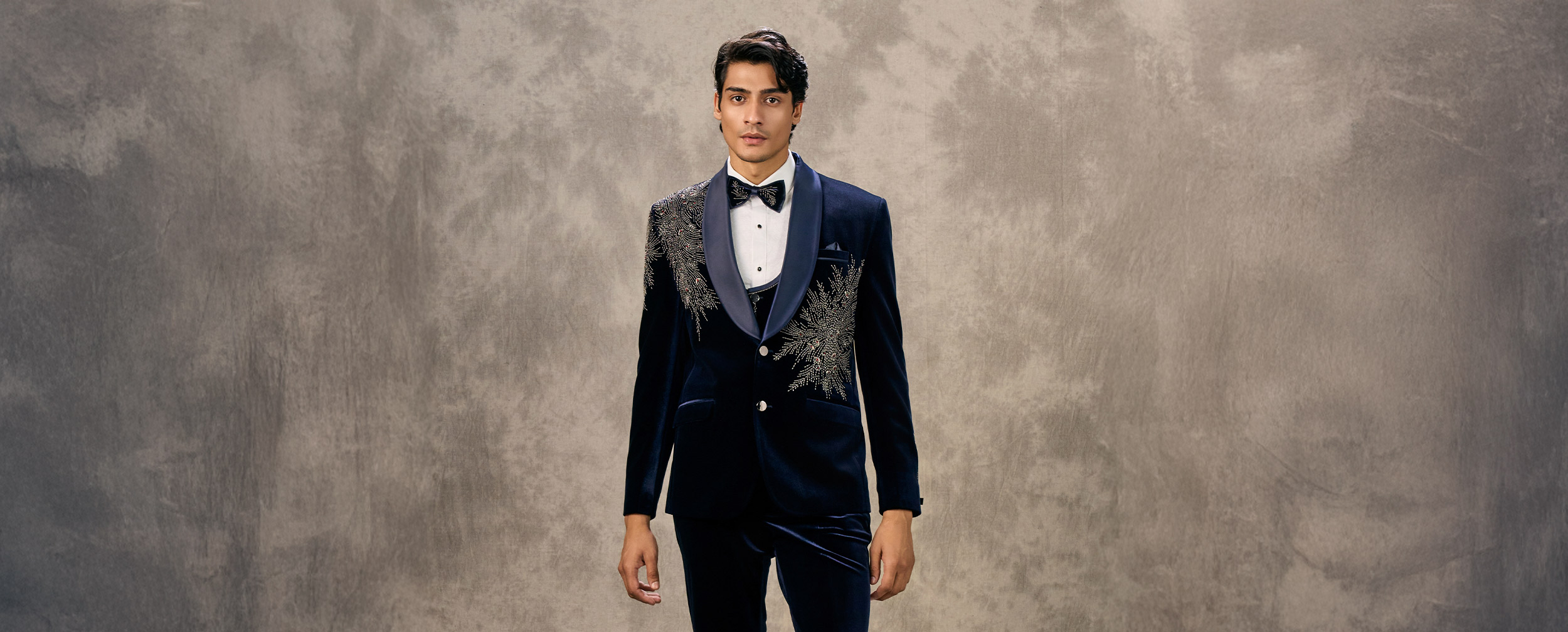 Man in a navy blue velvet tuxedo jacket with intricate silver embroidery and a black bow tie.